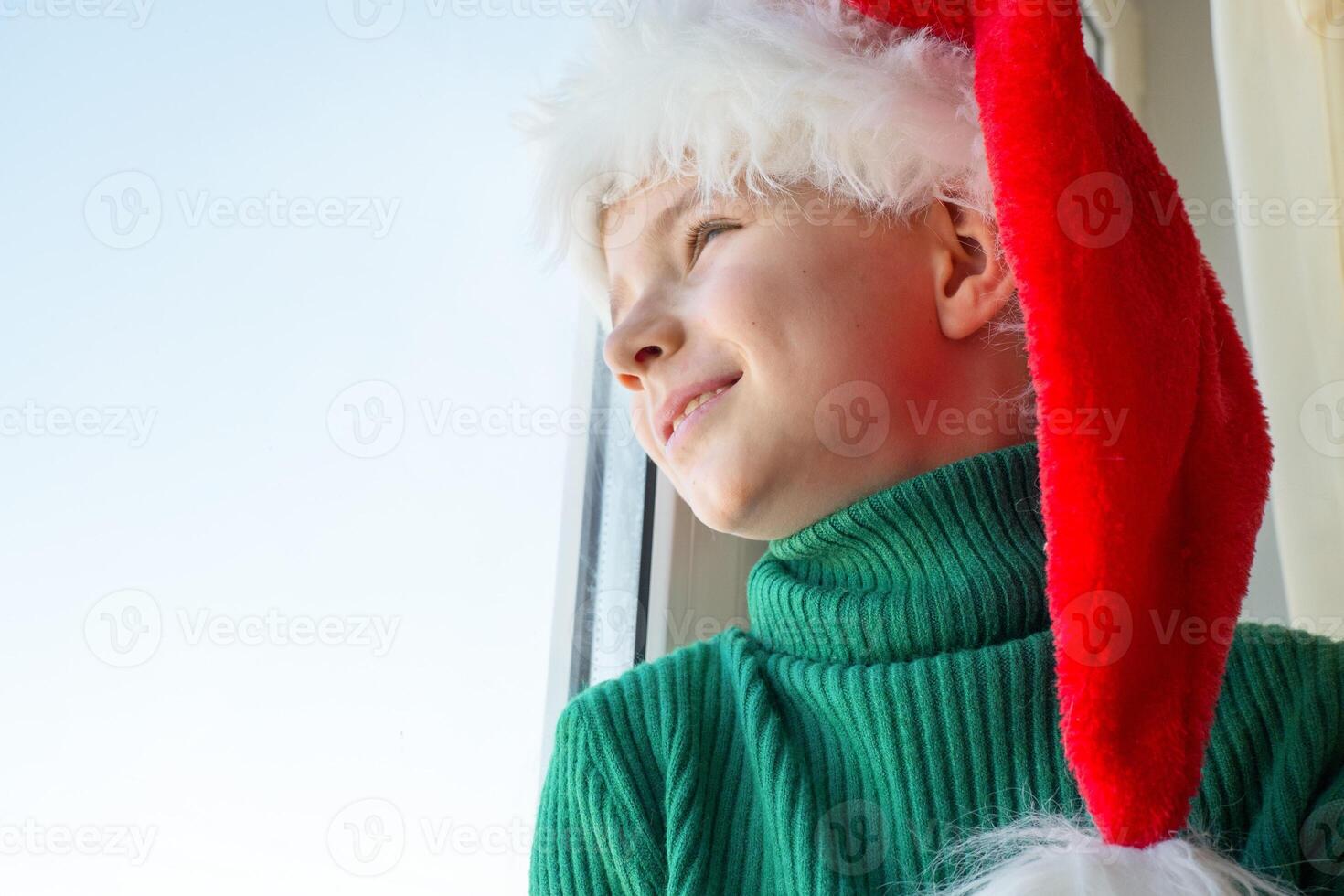 Children's dreams concept. Friendship. A lonely little boy in a Santa Claus hat is sad at the window waiting for gifts, looking out the window, the child is waiting for Santa Claus. photo