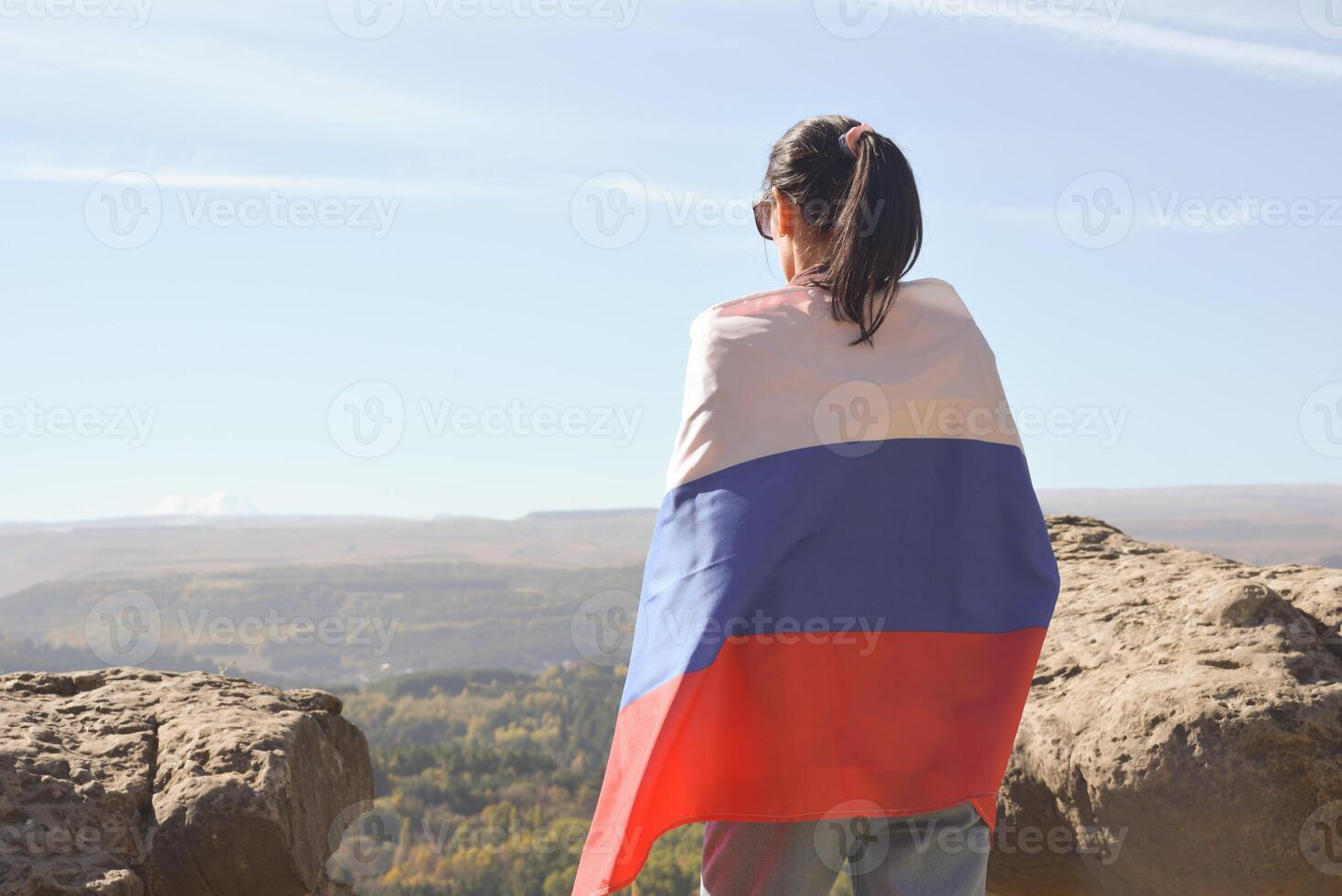 A girl tourist with a Russian flag on her shoulders stands on the top of a mountain and enjoys the beautiful views of the Caucasus Mountains. photo