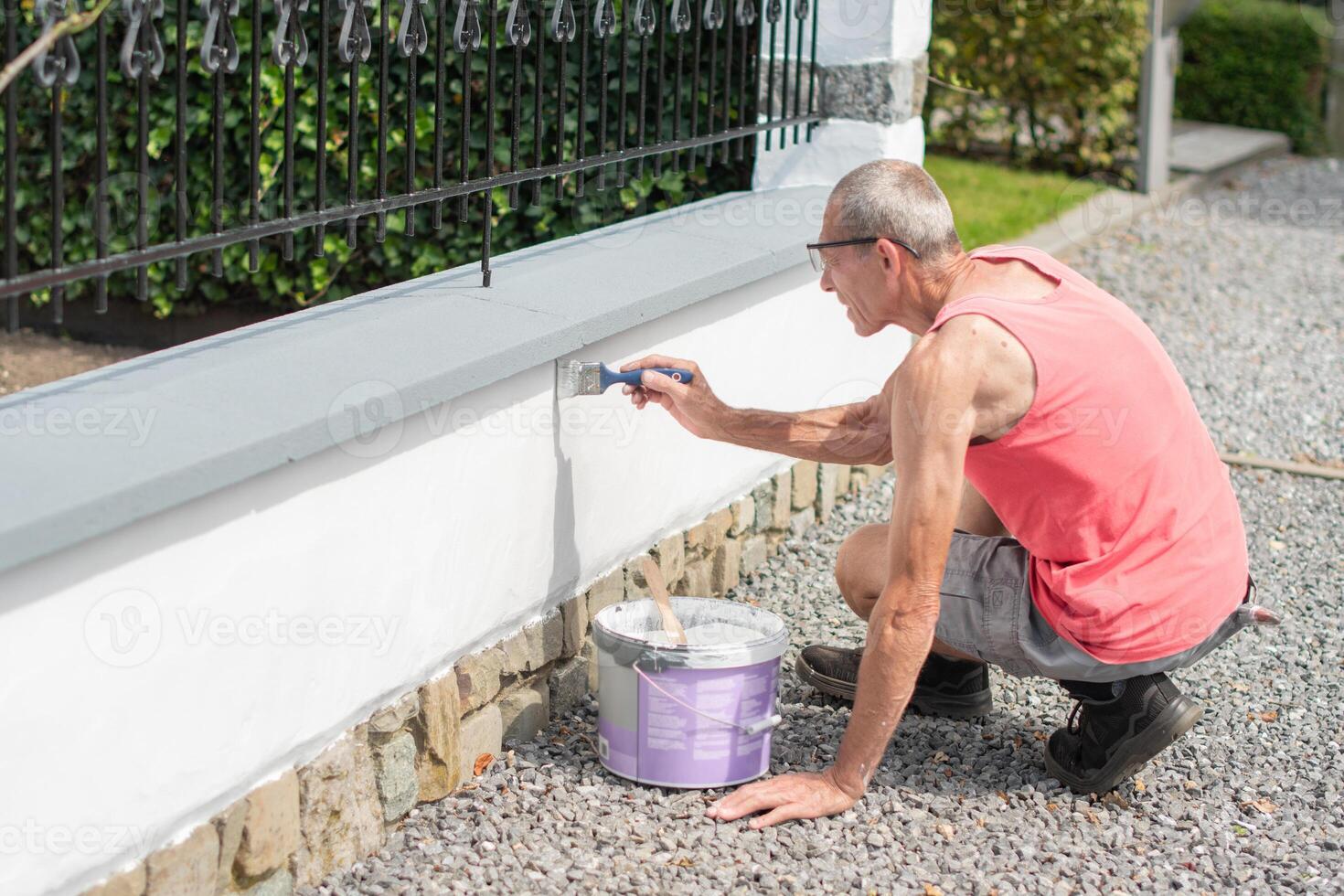 A middle-aged man paints a fence with white paint with a brush on a sunny summer day, repairs the damaged surface photo