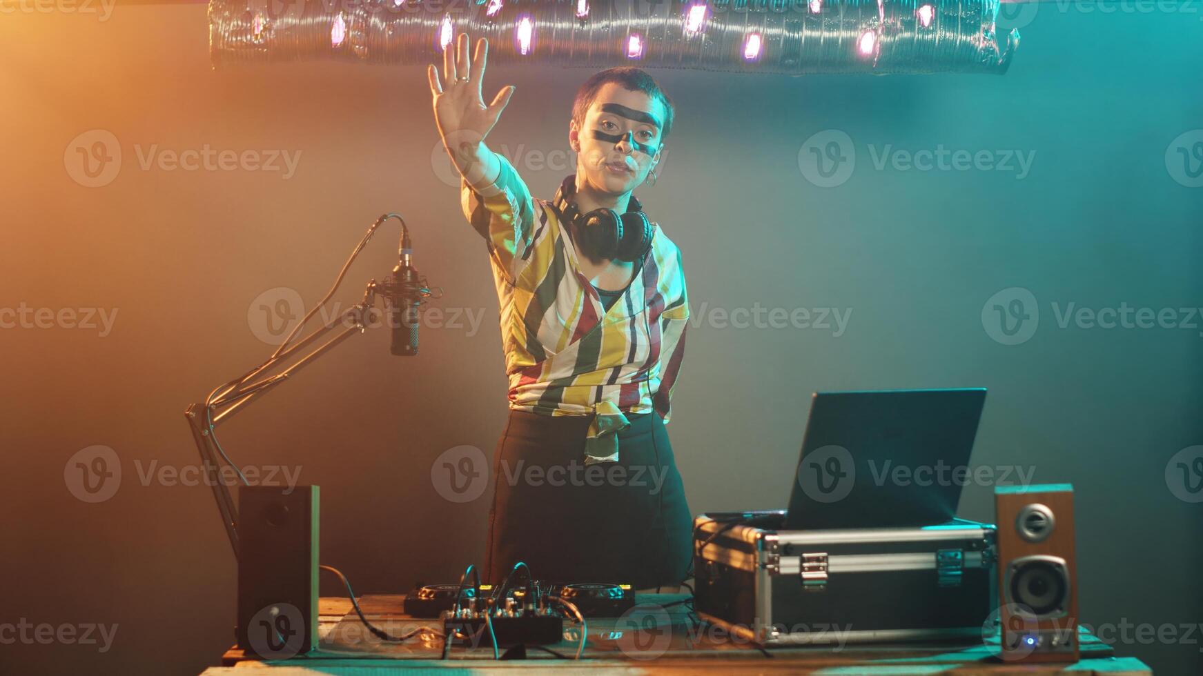 Funky performer at club raising palm to express stop sign on stage, trying to mix techno records and tunes on turntables. Woman artist using DJ instruments, adjusting bass control key. Tripod shot. photo