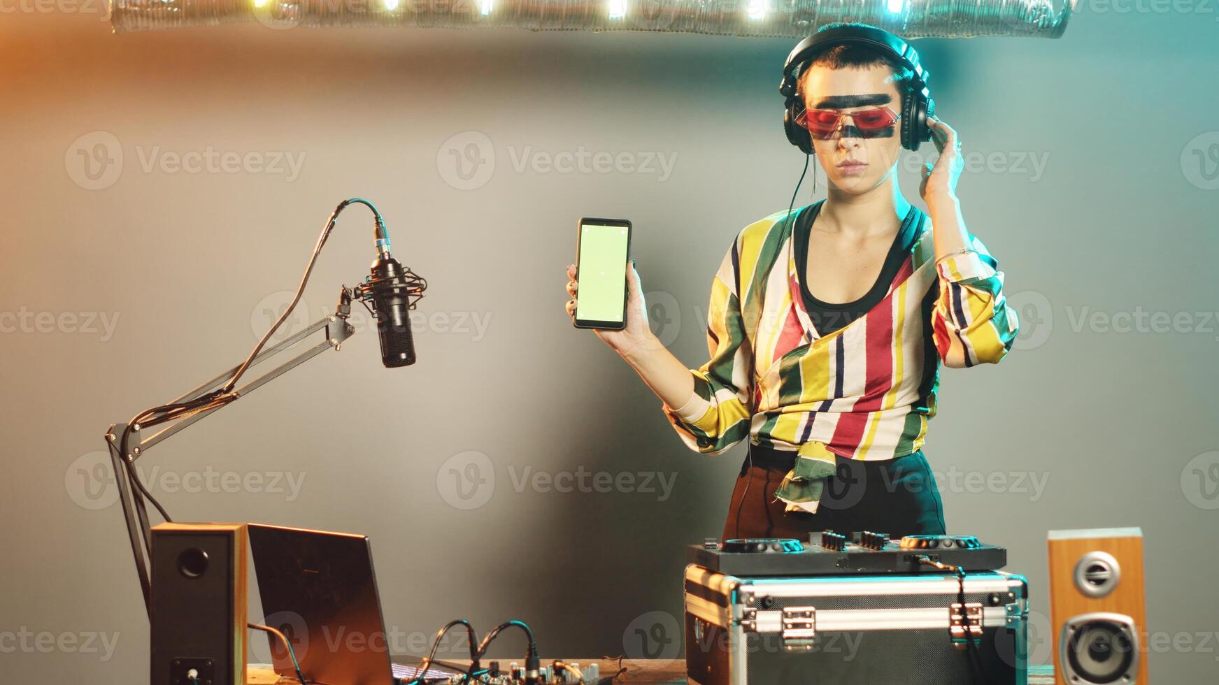 DJ performer points at greenscreen display while she uses mixer control keys for bass and volume, creating modern techno performance at club party. Woman with chromakey template. photo