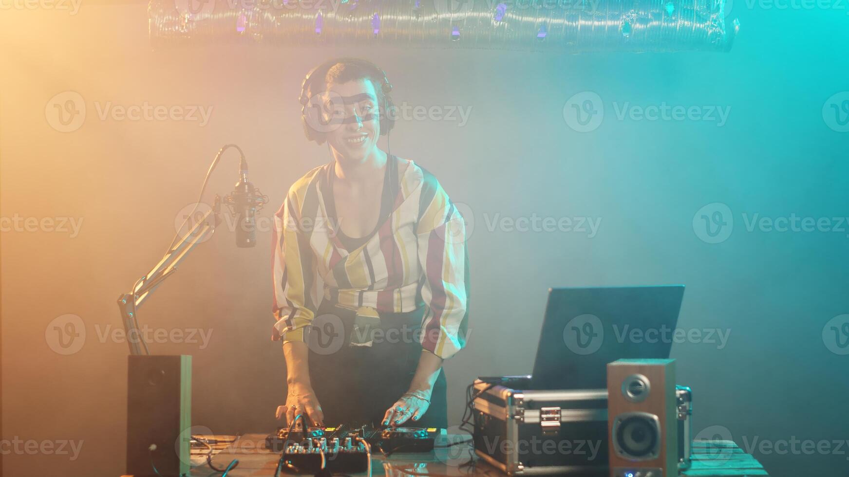 Female DJ playing techno music at stereo mixer, having fun with turntables instrument as disc jockey and mixing records at nightclub. Using headphones to test bass control key. Tripod shot. photo