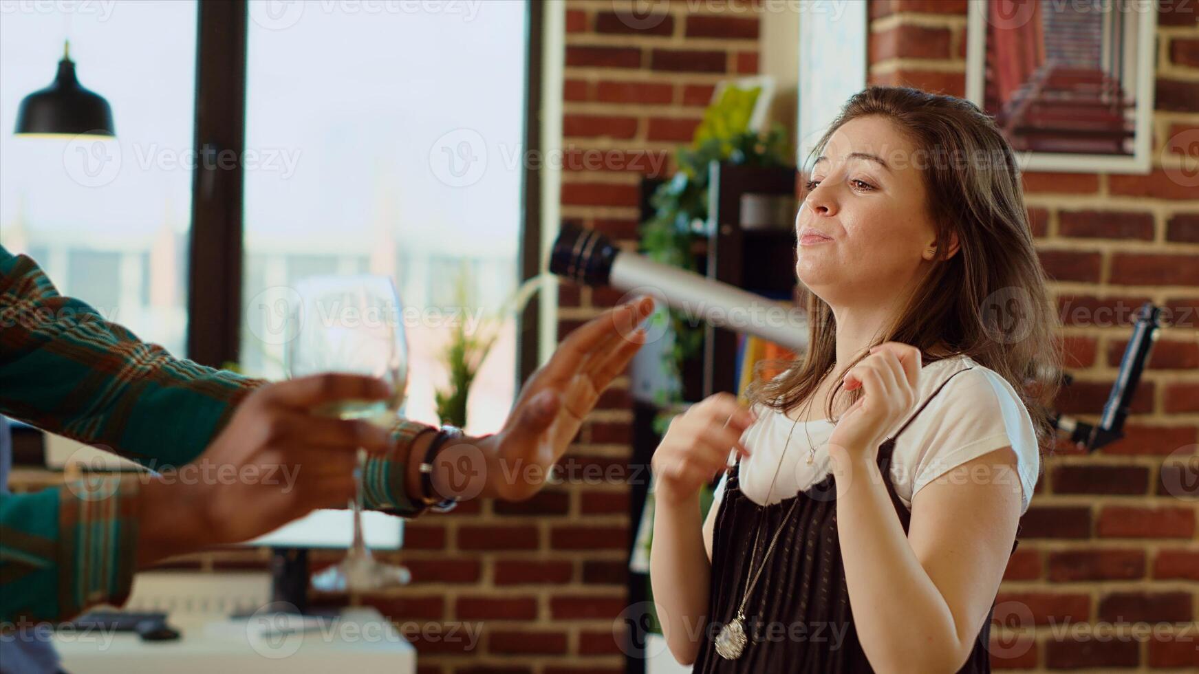 Joyful friends dancing at house party while drinking wine glasses, enjoying celebration. Multiracial group of people having fun indoors, doing entertaining dance moves to music in modern apartment photo