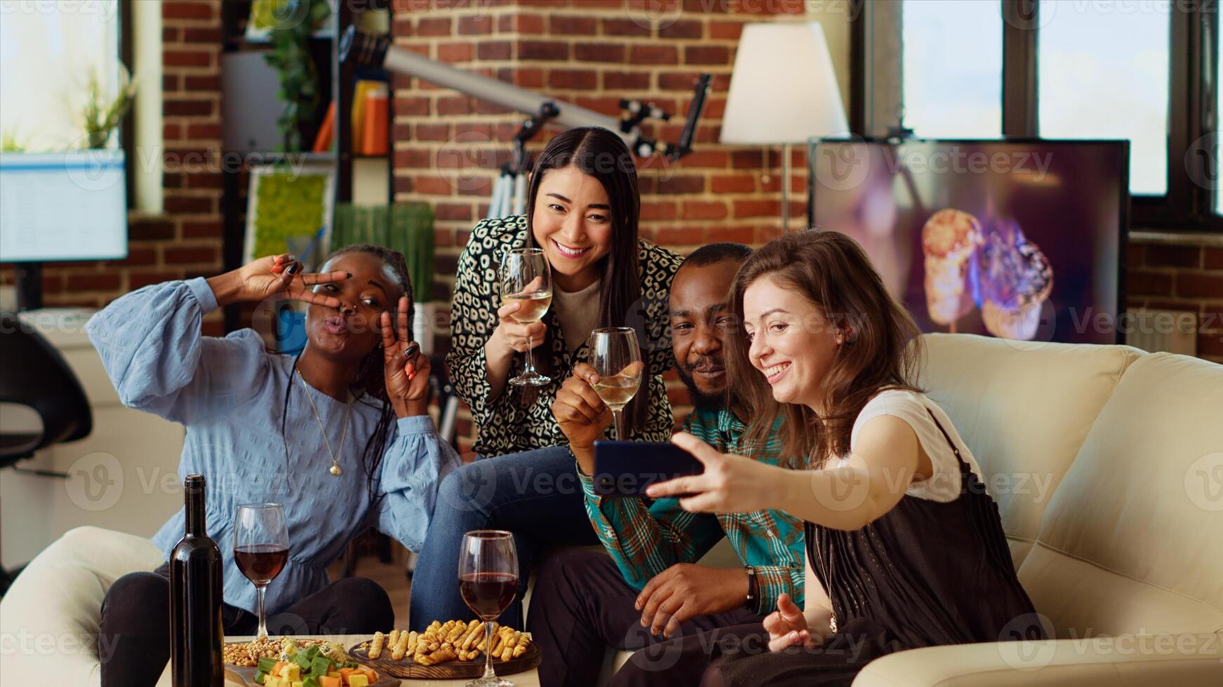 BIPOC friends taking group pictures with selfie camera smartphone at apartment party. Joyful mates in modern home posing for photo with wine and champagne glasses, making gestures