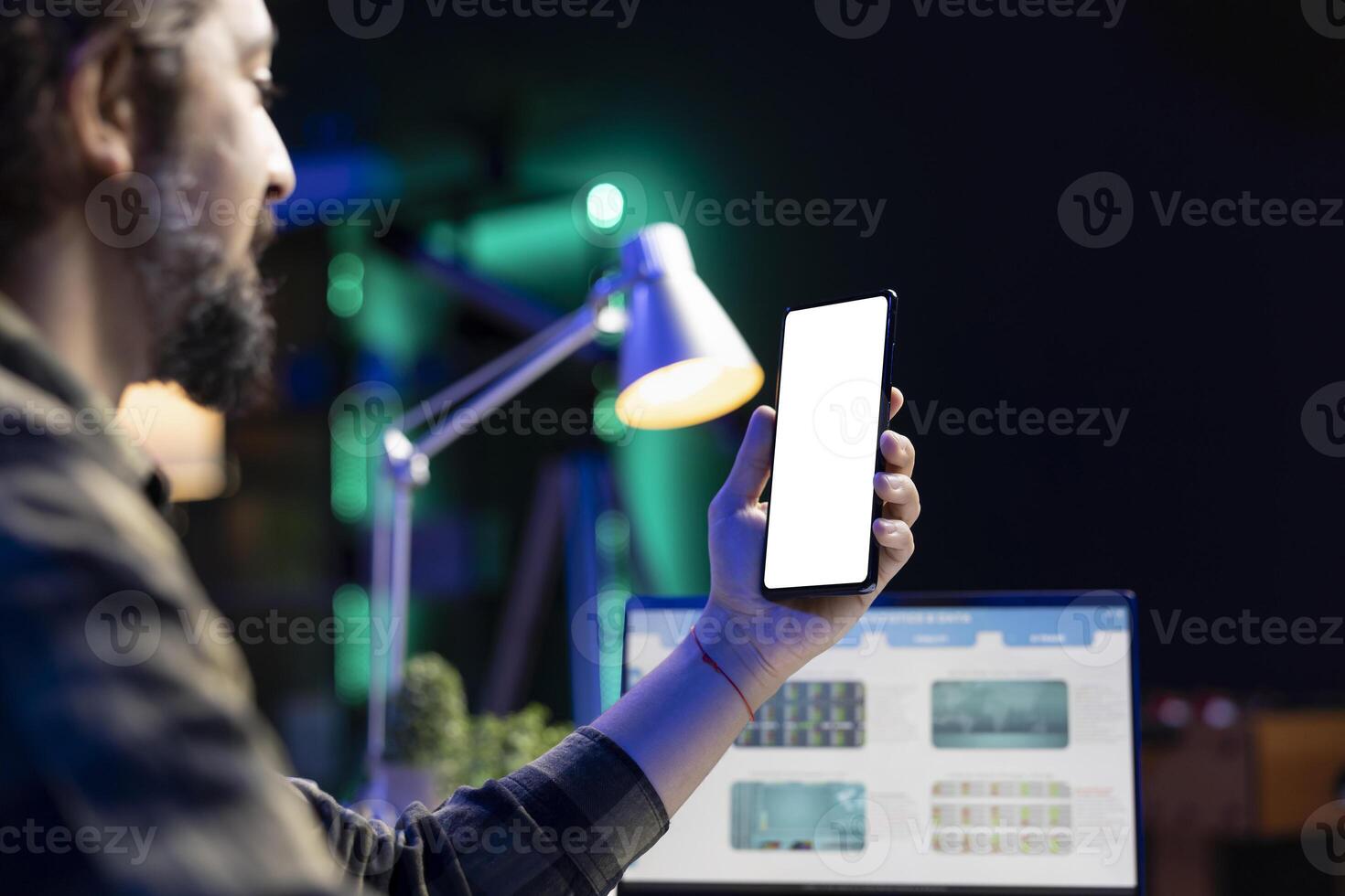Bearded man using cell phone with isolated white screen while using his laptop. Male freelancer holding smartphone with a blank chromakey template while working at his table. photo