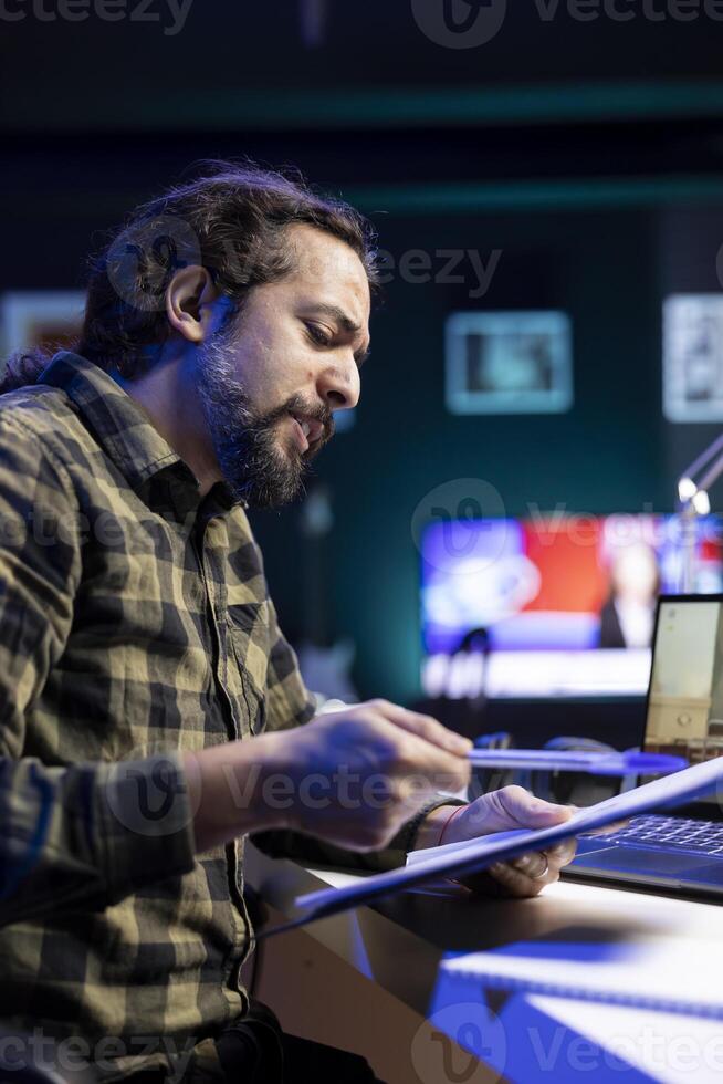 Male freelancer seated at the desk, going through his documents and discussing in a virtual meeting on his laptop. Young man reviewing information on clipboard and using his portable computer. photo