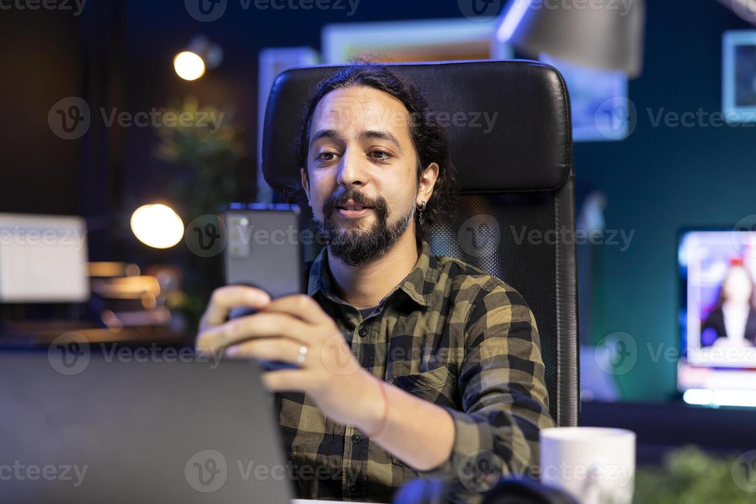 Bearded man sits in front of a laptop, holding his cell phone and smiling while browsing the internet. A positive young male uses his mobile device to check his social media accounts. photo