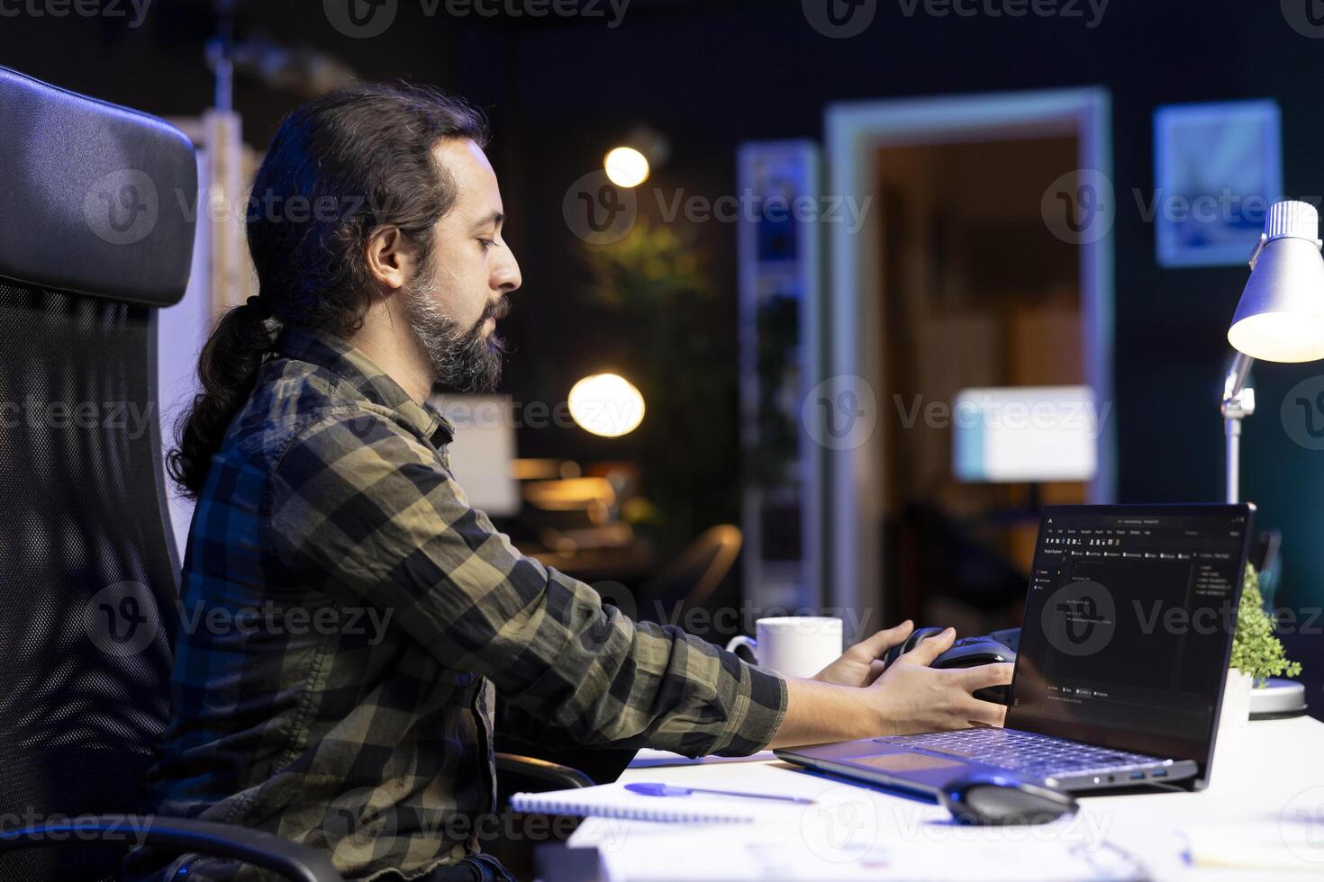 Male individual taking his wireless headset from table for music while working on his laptop. Bearded man touching his headphones while his device screen displays software codes. photo