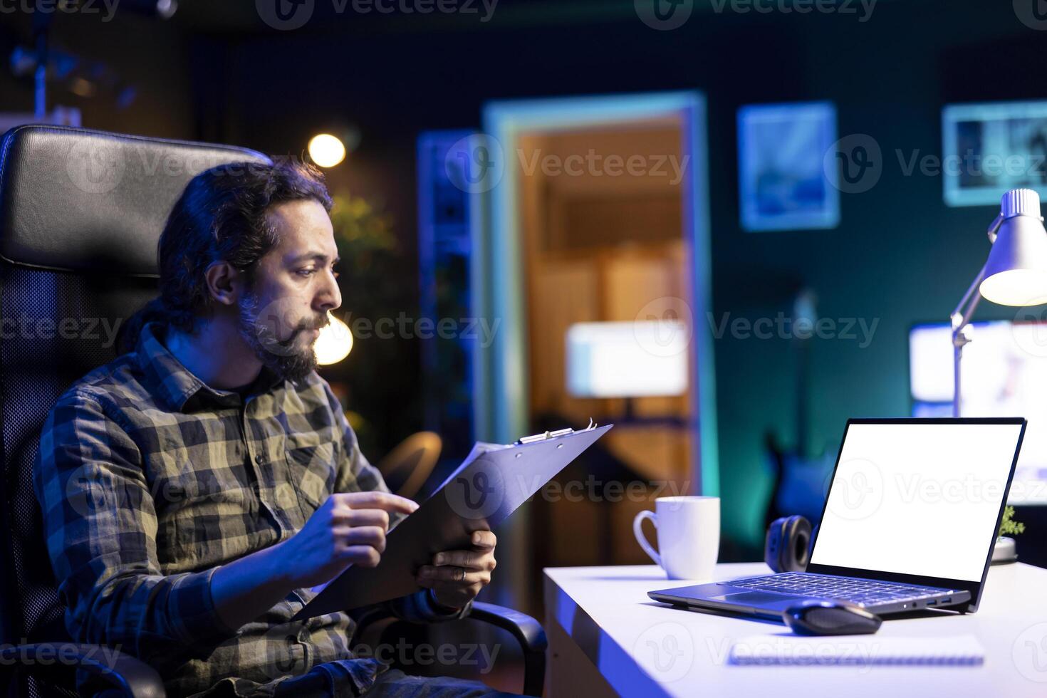 Bearded man is utilizing his digital laptop which is showing a blank chromakey template. Male individual holding clipboard and looking at his device which has an isolated mockup display. photo