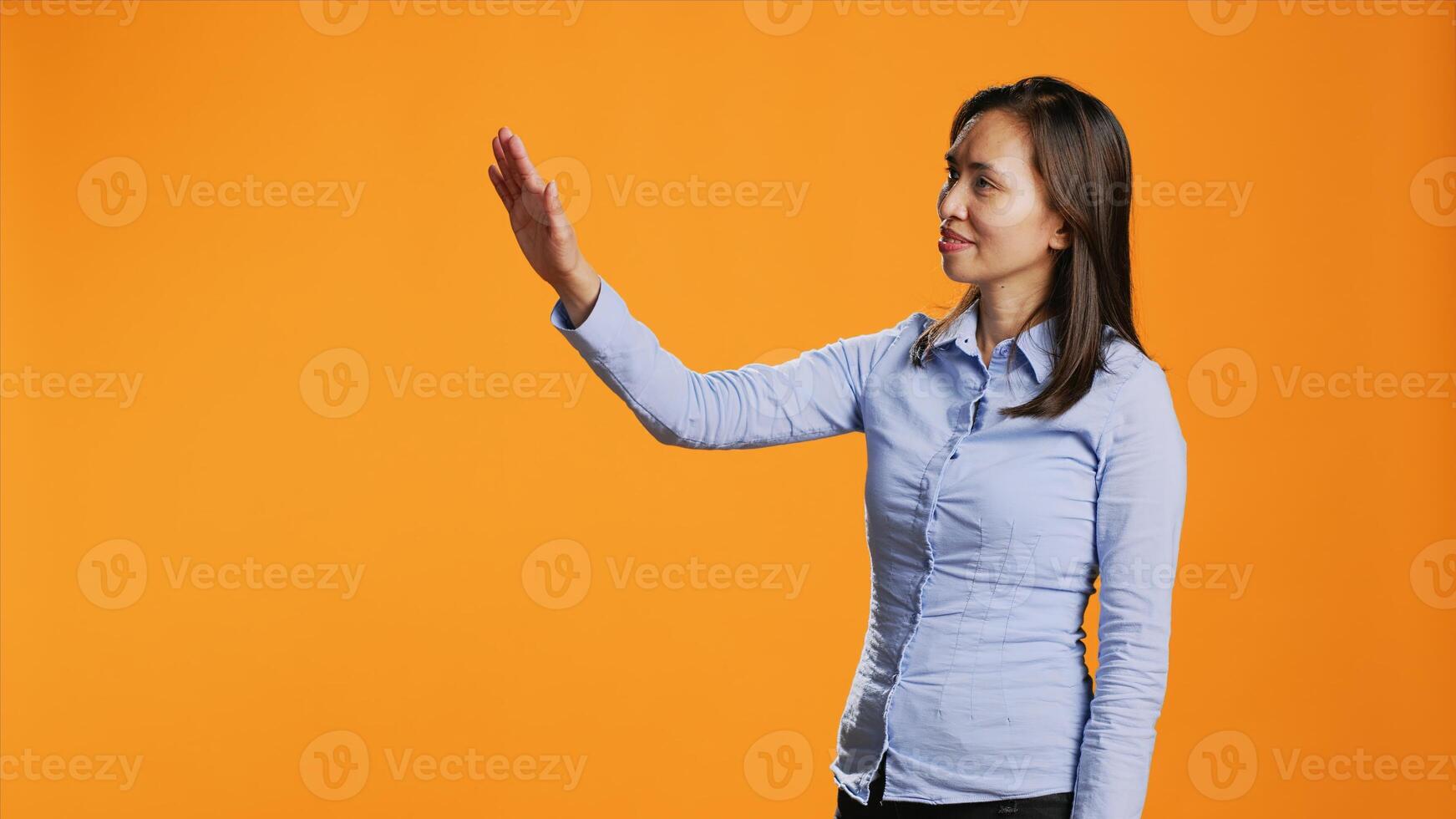 Asian woman examines holographic image reflection in studio, looking at modern artificial intelligence hologram over orange background. Confident person using metaverse projection. photo