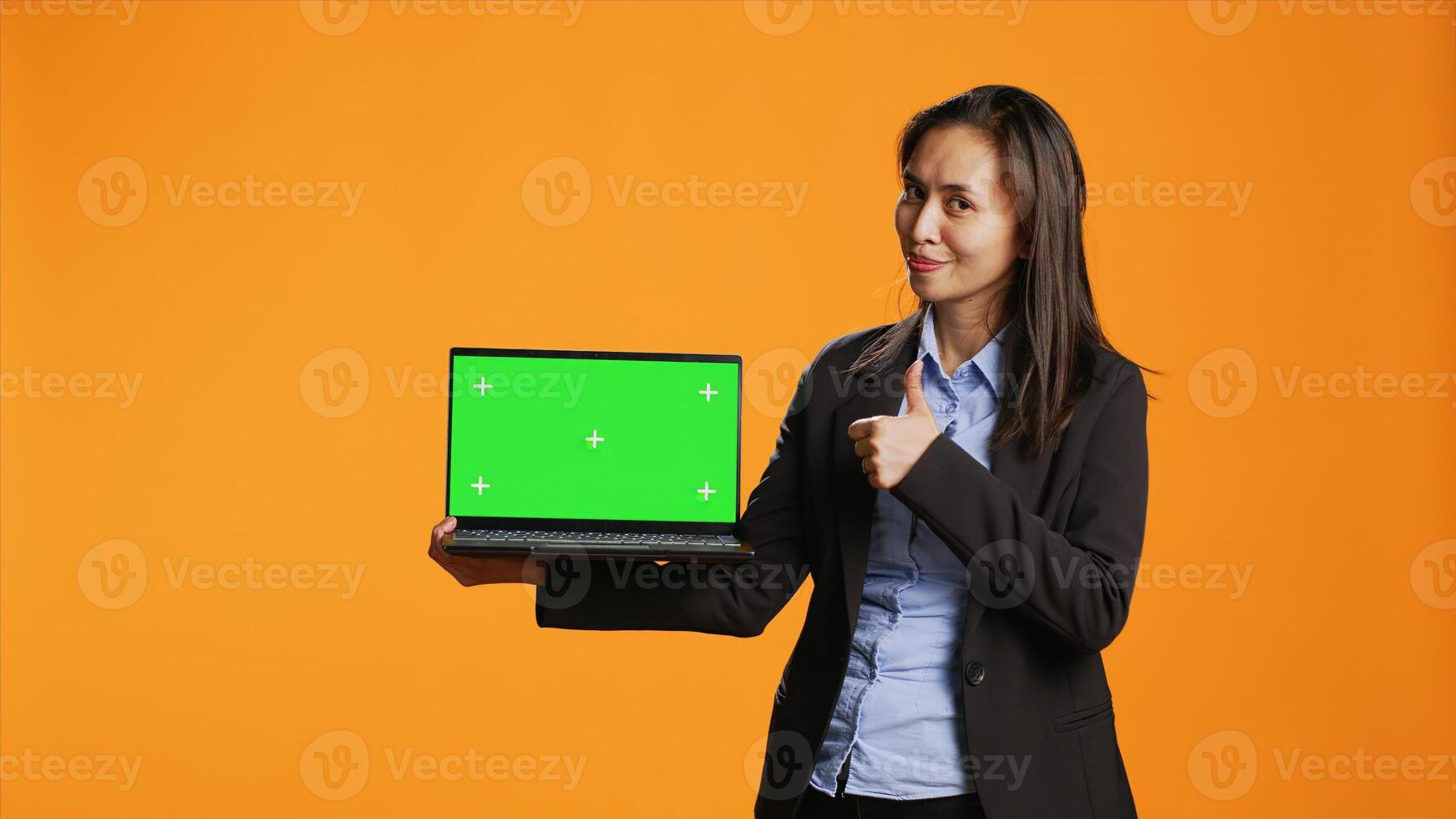 Business person holds laptop with greenscreen on camera, showing blank copyspace display on portable pc. Asian woman in formal suit presents computer with chromakey screen in studio. photo