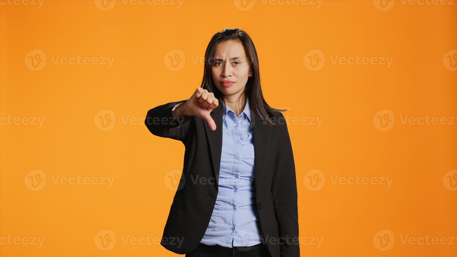 Asian female model giving dislike in front of camera, showing thumbs down with displeased facial expressions. Businesswoman does rejection and disapproval gesture over backdrop. photo