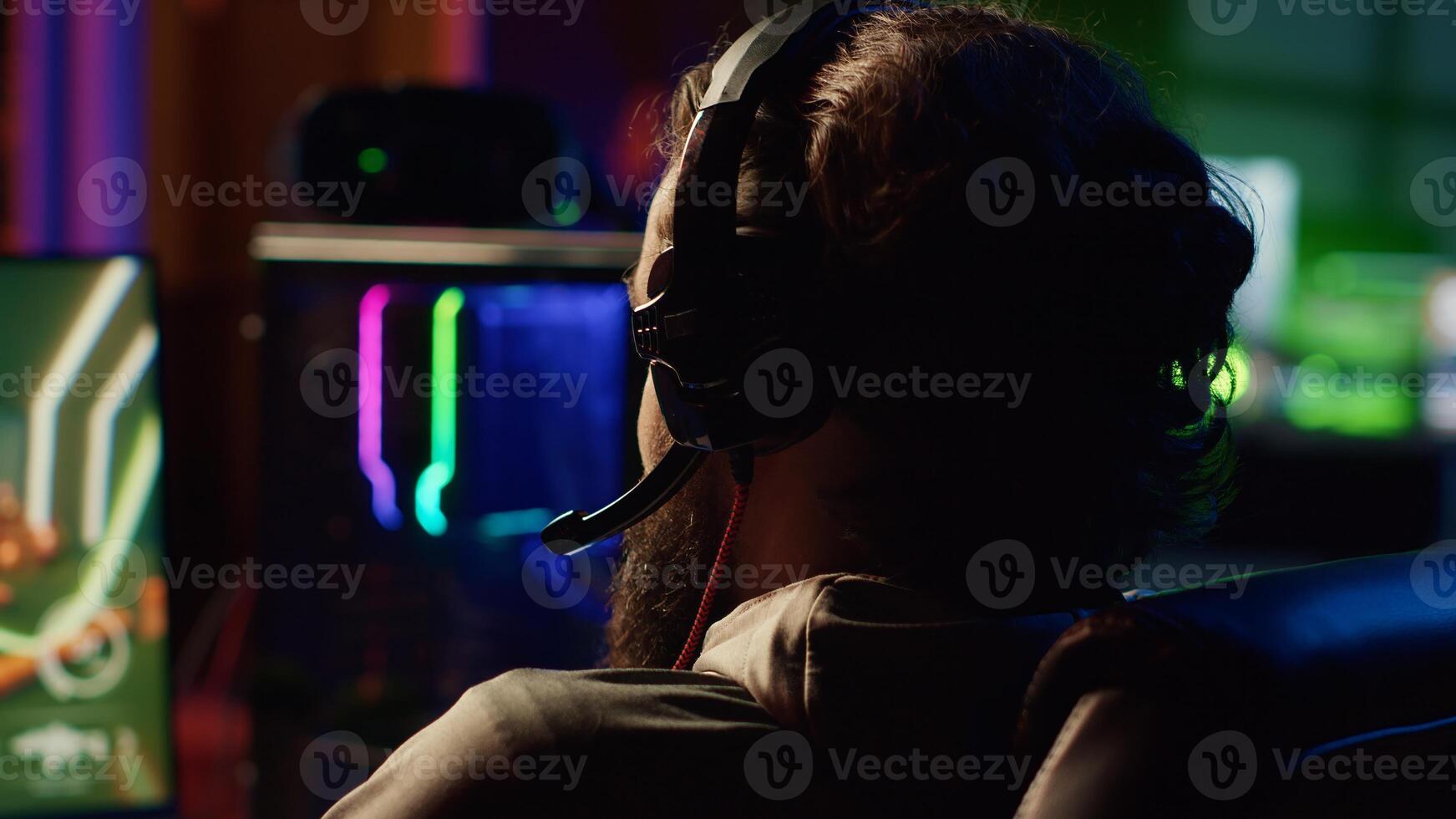 Gamer using headphones mic to talk with online friends from his dark living room, playing engaging video games on gaming PC at computer desk, relaxing after work, close up shot photo