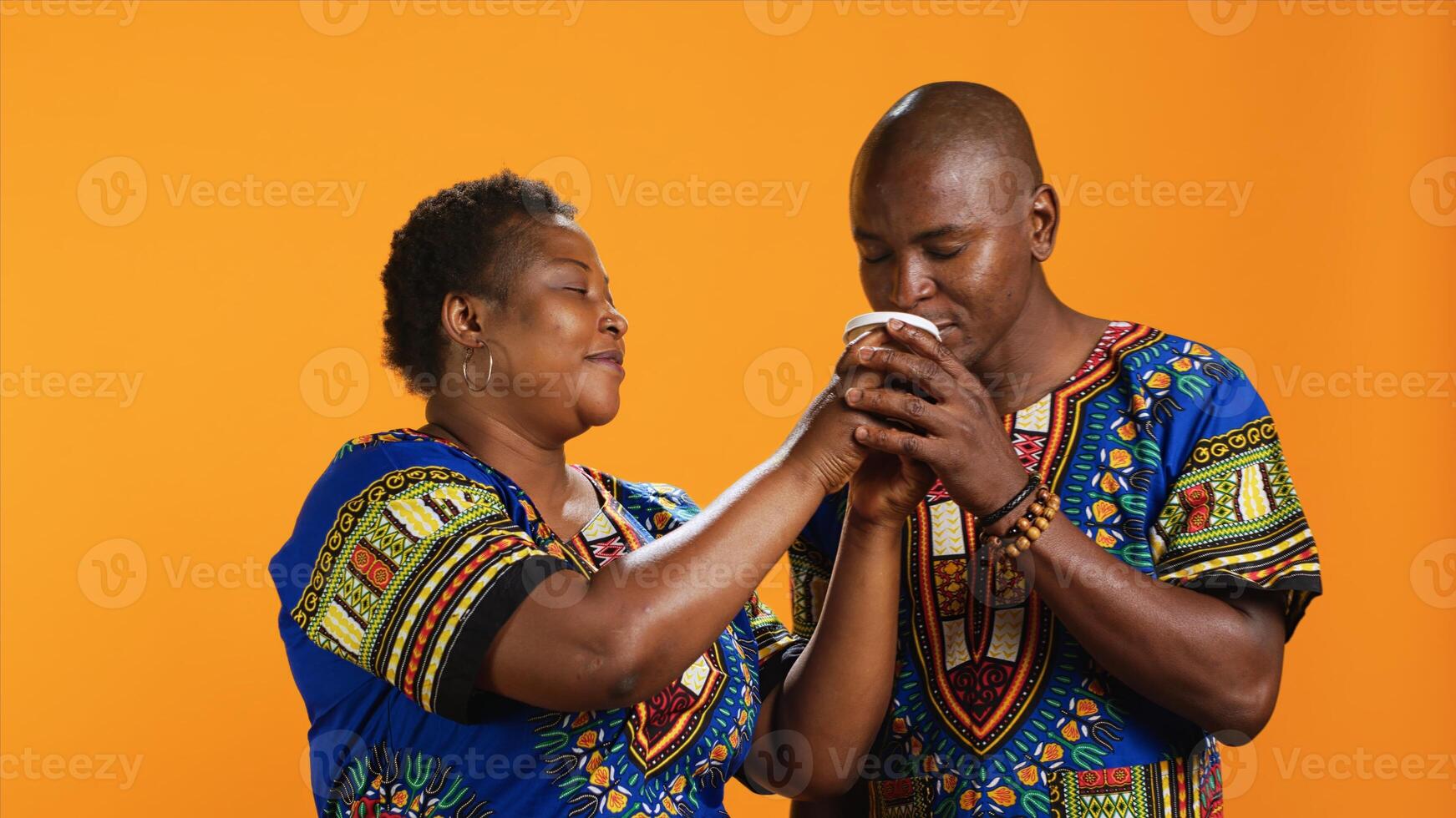 Ethnic couple enjoying aroma of fresh coffee drink, smelling caffeine beverage while they pose over orange background. Young partners in traditional attire loving scent of refreshment. photo