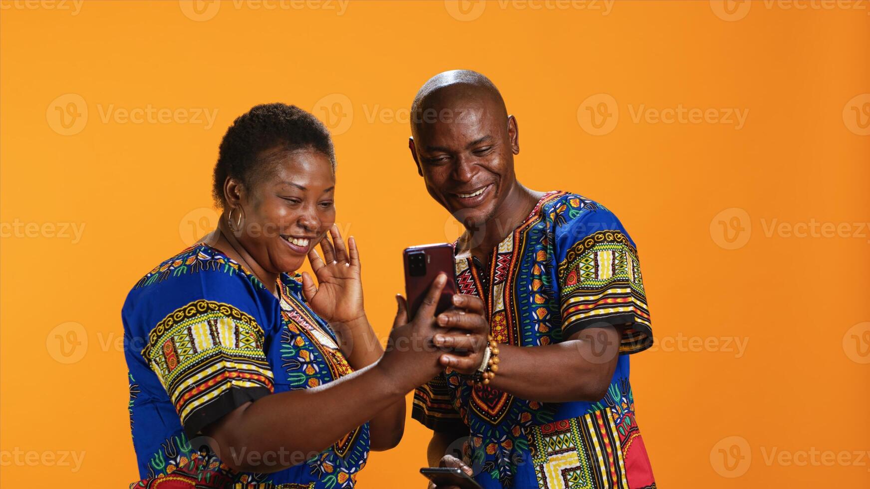 Traditional african american couple browsing websites on smartphone, checking photos on social media app and laughing at memes. Joyful people in colorful attire looking at pictures online.