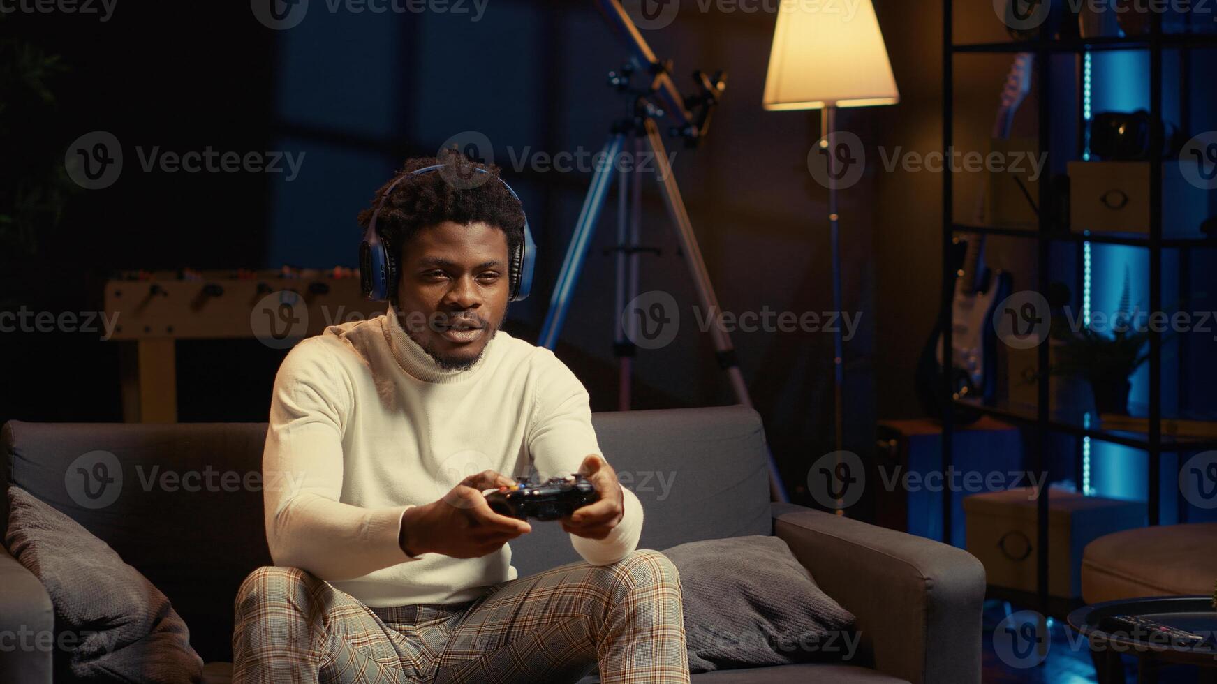 African american man in living room playing video games on gaming console, smashing controller buttons. Gamer wearing headphones, battling enemies in online multiplayer shooter from cozy apartment photo