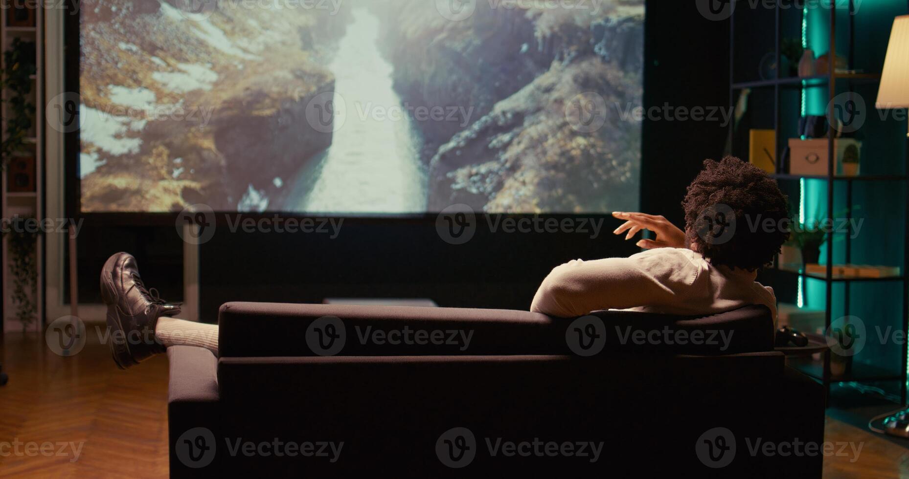 African american man watching nature documentary with beautiful cinematography on video projector. Person enjoying mountains and rivers scenery shots in home theatre, relaxing on couch photo