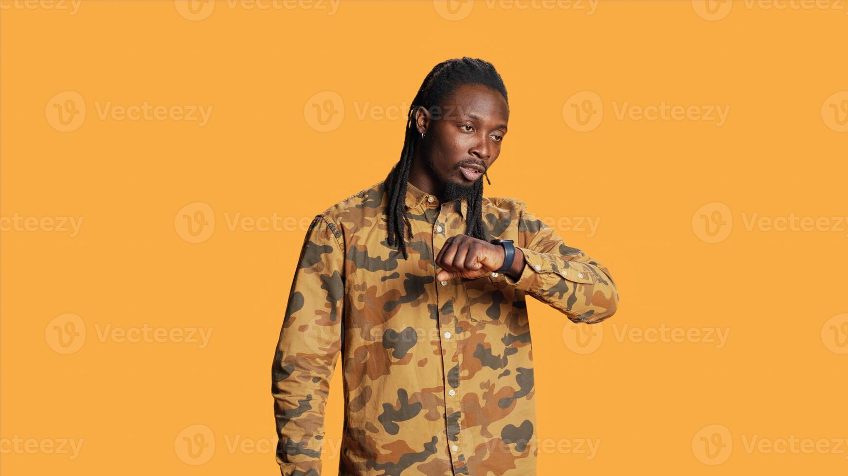 Modern african american guy takes call on smartwatch in studio, having remote conversation on telephone line using wristwatch over orange background. Smiling cheerful person in discussion. photo