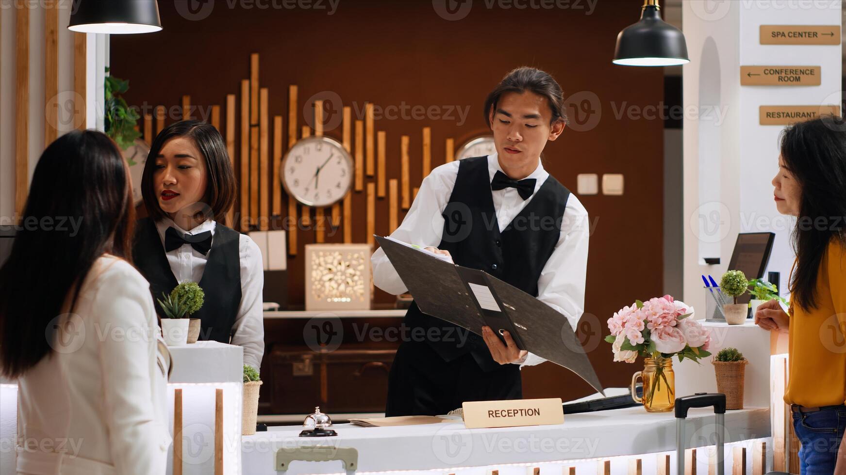 Male receptionist searching guest reservation in the registry, greeting asian woman at reception front desk. Employee helping tourist with registration process, hospitality industry. Handheld shot. photo