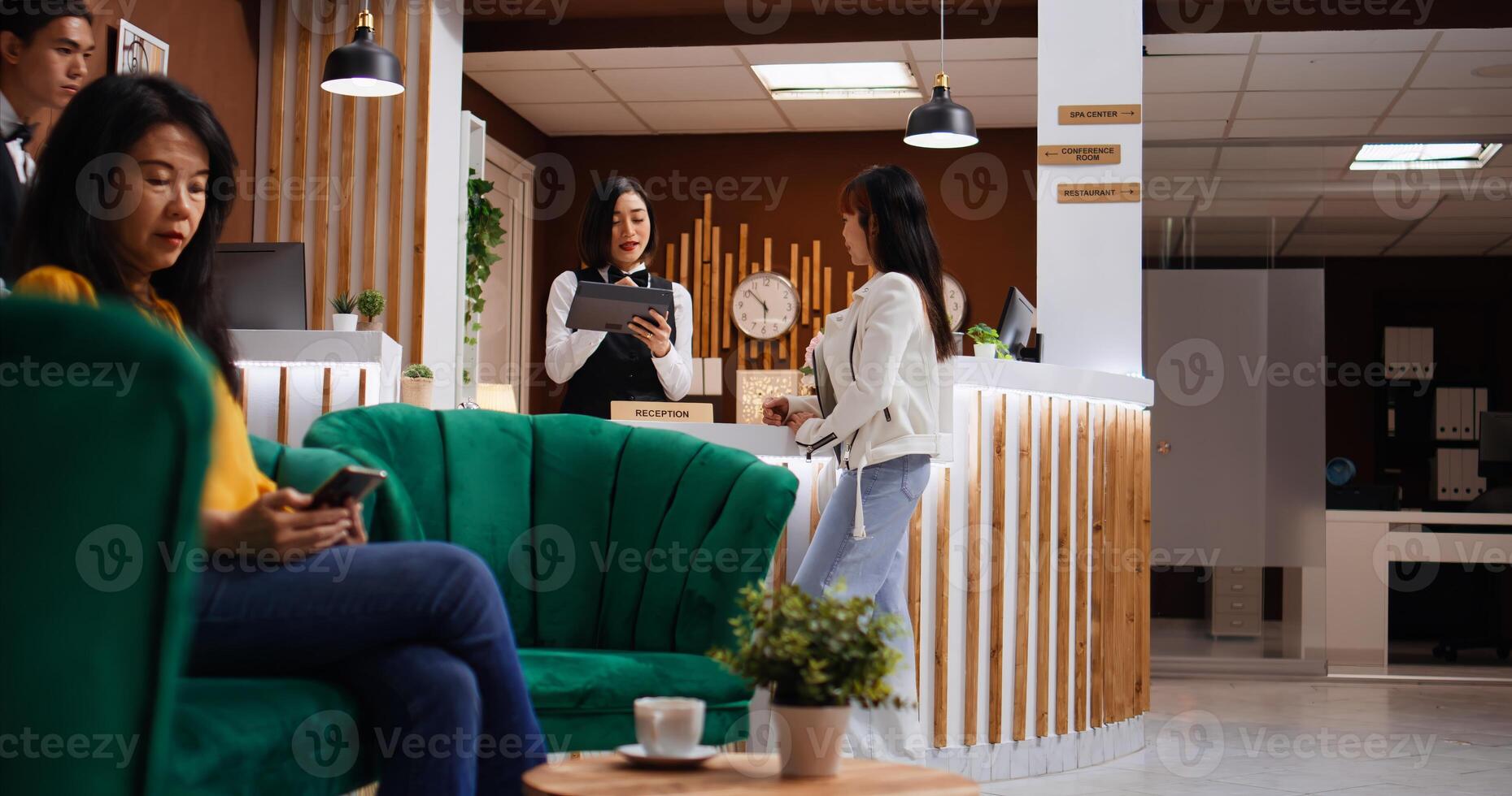 Asian guest signing check in papers on tablet at reception, registering her room confirmation while bellboy is helping with her luggage. Five star hotel staff team friendly welcoming client. photo