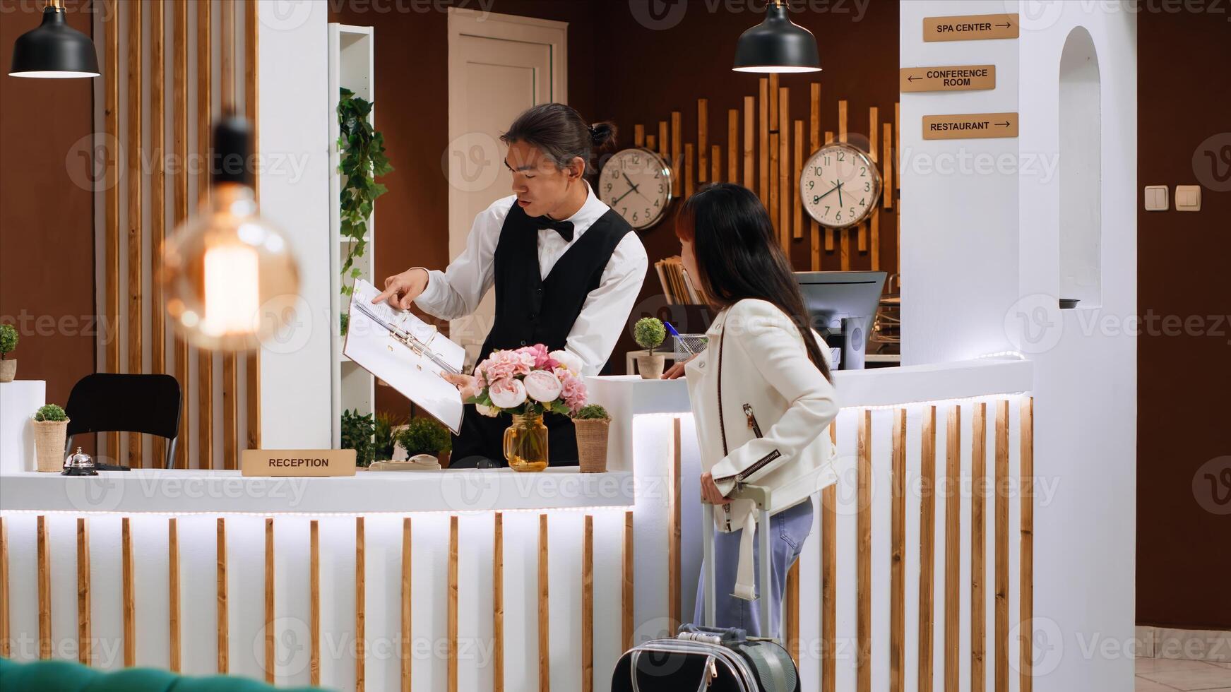 Asian traveler asking front desk staff about facilities after arriving at luxurious holiday resort, showing her room booking to the employee. Young man greeting client in reception lobby. photo