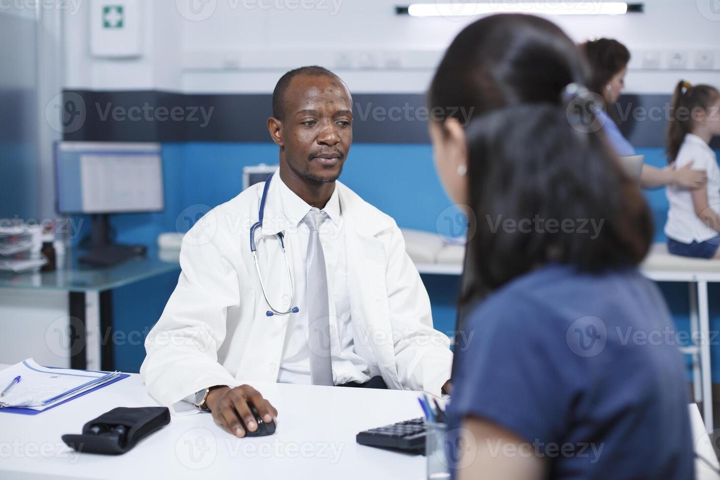 Selective focus on the african american doctor using the computer while speaking with the caucasian female patient in the clinic. The atmosphere is professional and reassuring. photo
