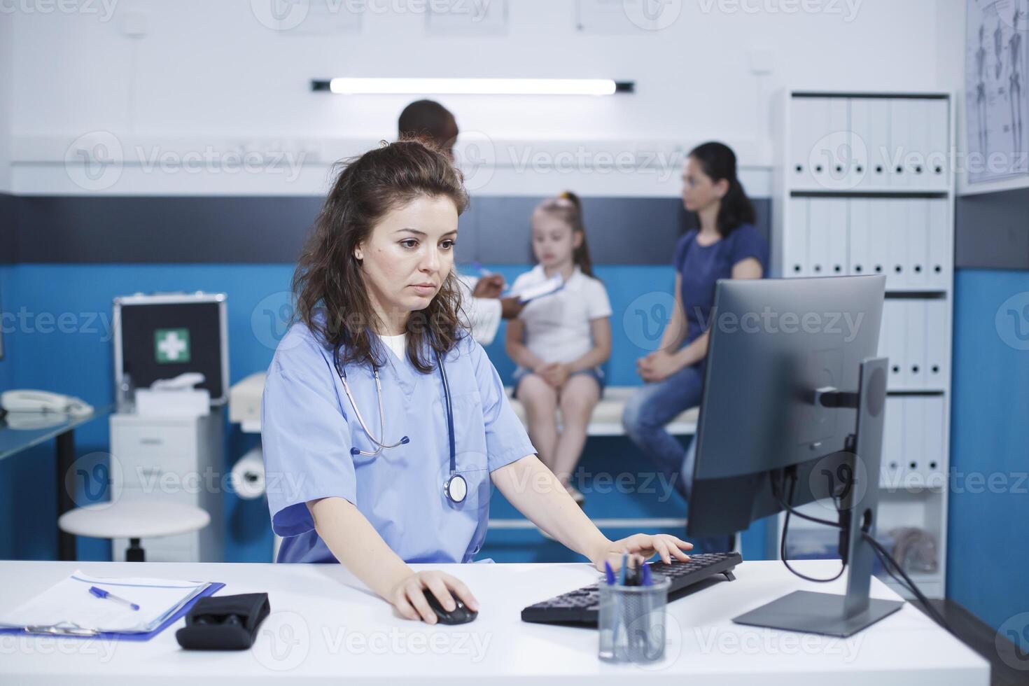 Caucasian female nurse using the computer desktop, to verify patient information and appointments, while male doctor assists mother and daughter. A scene of healthcare and communication. photo