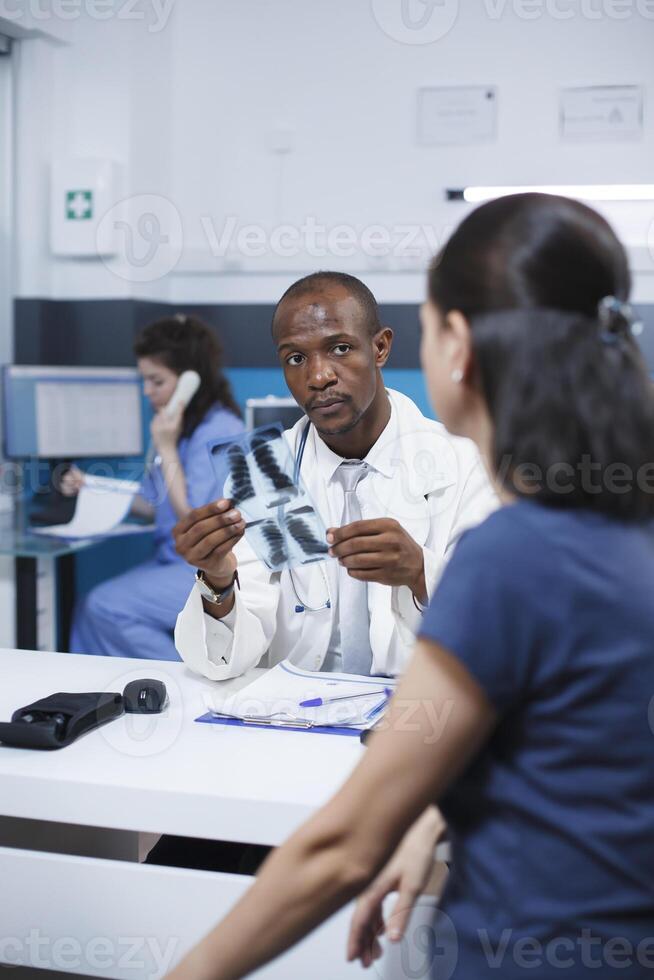 African American female specialist reviewing x-ray image and analyzing imaging results. Radiology expert examines radiography scan and provides diagnostic results to caucasian woman patient. photo