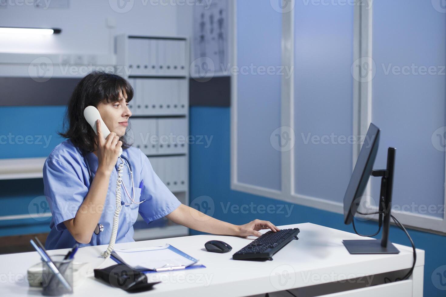 In the hospital office, a female medical professional takes calls and sets up appointments. Nurse utilizes a telephone at a desk in a contemporary clinic while focusing on a pc monitor. photo