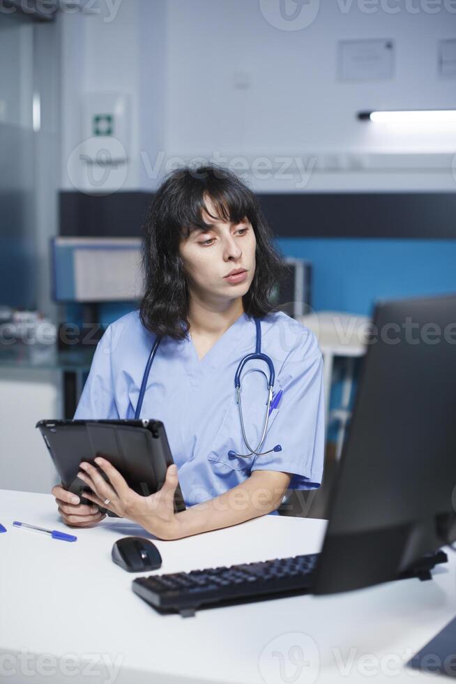For healthcare, a medical assistant uses a computer and tablet with a touch screen. Working late at night, a female nurse holds a gadget and reviews patient consultations. Expert in electronic devices photo