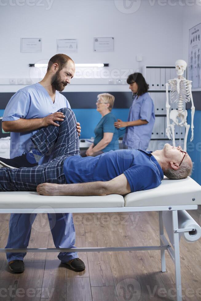 Physiotherapist is assisting elderly man recovering from a leg injury. Senior patient receiving help from a chiropractor for knee discomfort and muscular tightness to aid in rehabilitation. photo