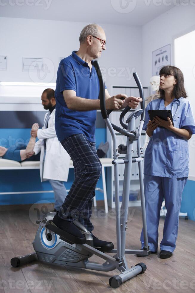 Elderly male patient undergoing physiotherapy on stationary bicycle, with assistance of nurse in blue scrubs. Medical assistant using electronic tablet assists senior citizen with physical therapy. photo