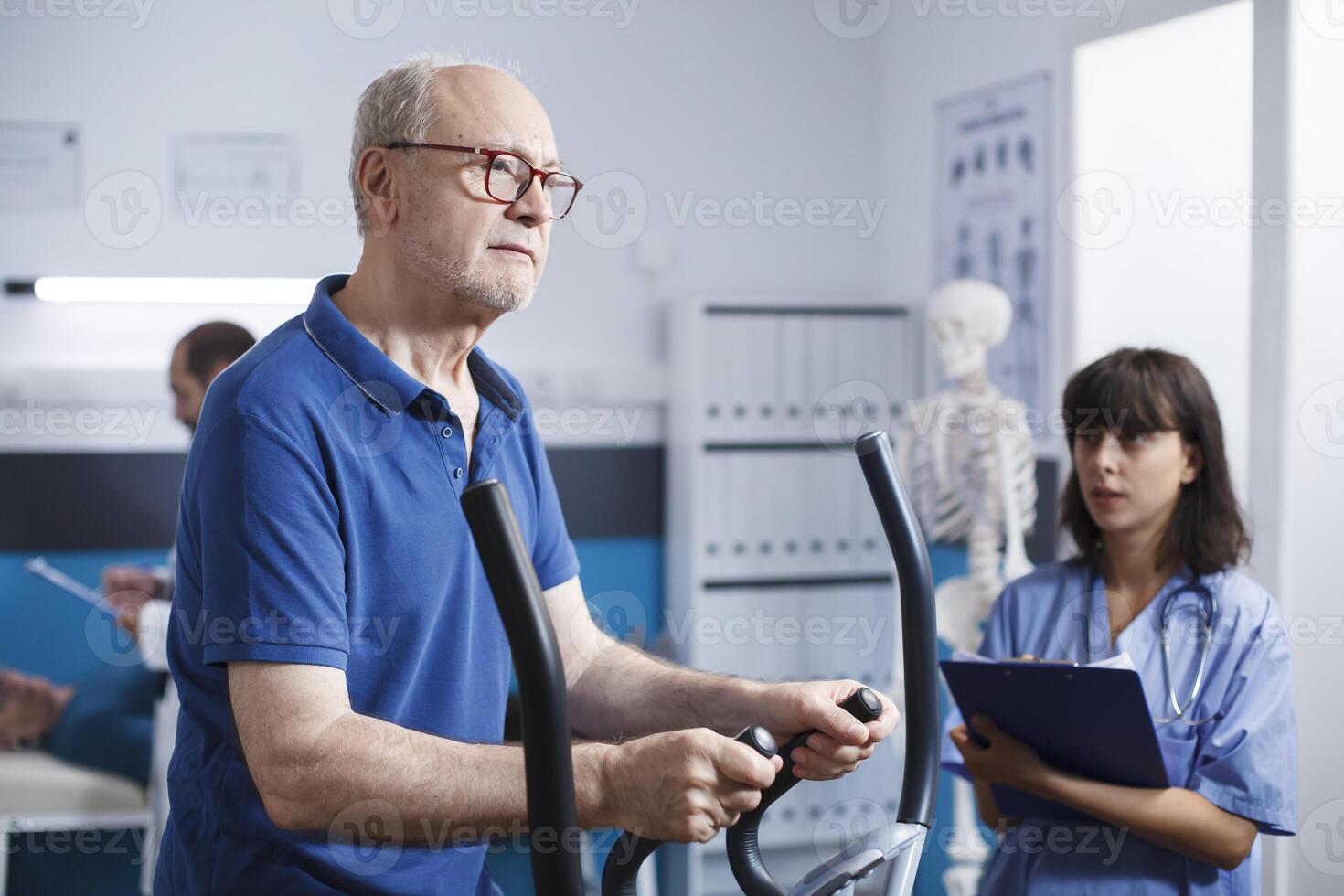 Caucasian old patient using stationary bicycle for physical therapy with help of female medical assistant in blue scrubs. Senior citizen in hospital for rehabilitation to heal muscle pain. photo