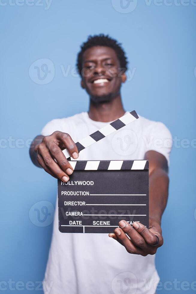 retrato de negro hombre utilizando un claqueta a podar secuencias en el película negocio. joven masculino adulto riendo y mostrando pizarron a cámara mientras participación cineasta objeto para rodaje. foto