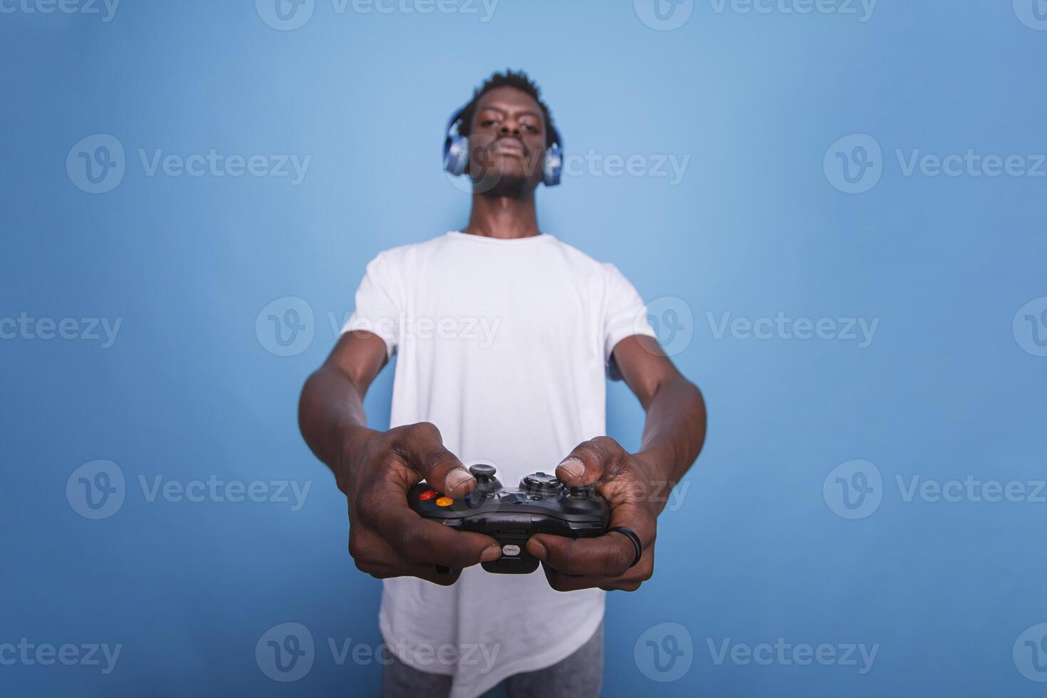africano americano vídeo jugador vistiendo auriculares y participación un palanca de mando hacia el cámara. juvenil negro hombre en pie en frente de azul antecedentes mientras utilizando un dispositivo para en línea juego de azar. foto