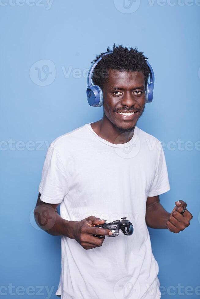 retrato de africano americano jugador celebrando victoria de vídeo juego en frente de aislado azul antecedentes. emocionado negro hombre con auriculares aplausos mientras participación un inalámbrico juego controlador. foto