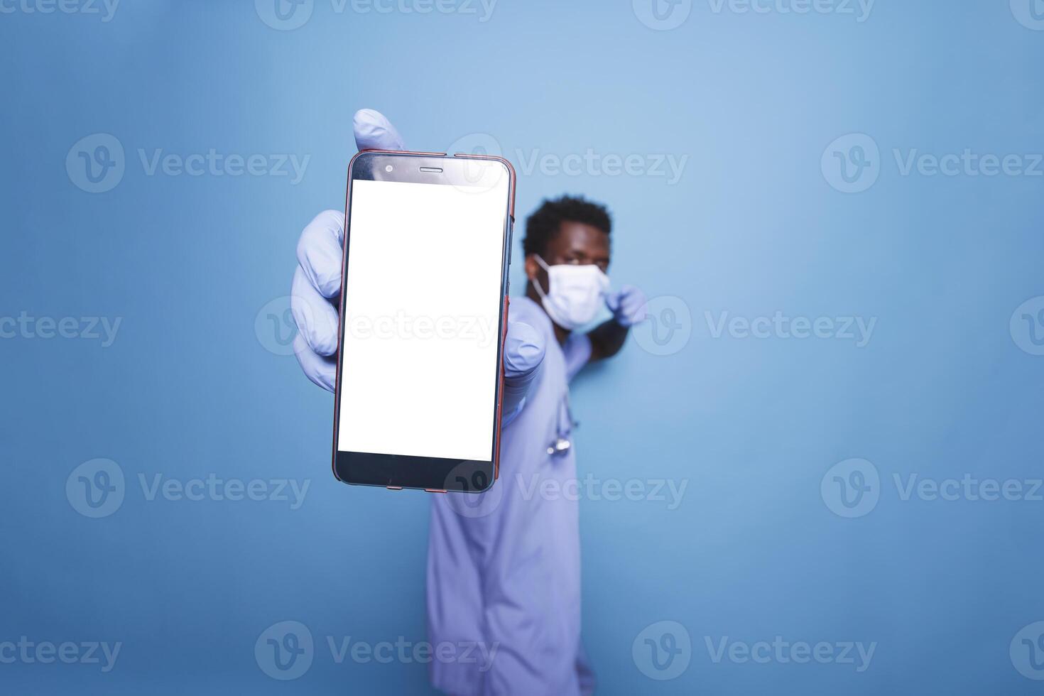 Nurse of African American ethnicity in scrubs and face mask holds a smartphone displaying blank chromakey mockup template. Black man grasps mobile device with white screen close to camera. photo