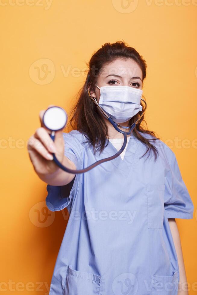 Close-up of female physician wearing blue scrubs grasping stethoscope in front of isolated background. Nurse with face mask gripping a medical instrument towards camera in studio. photo
