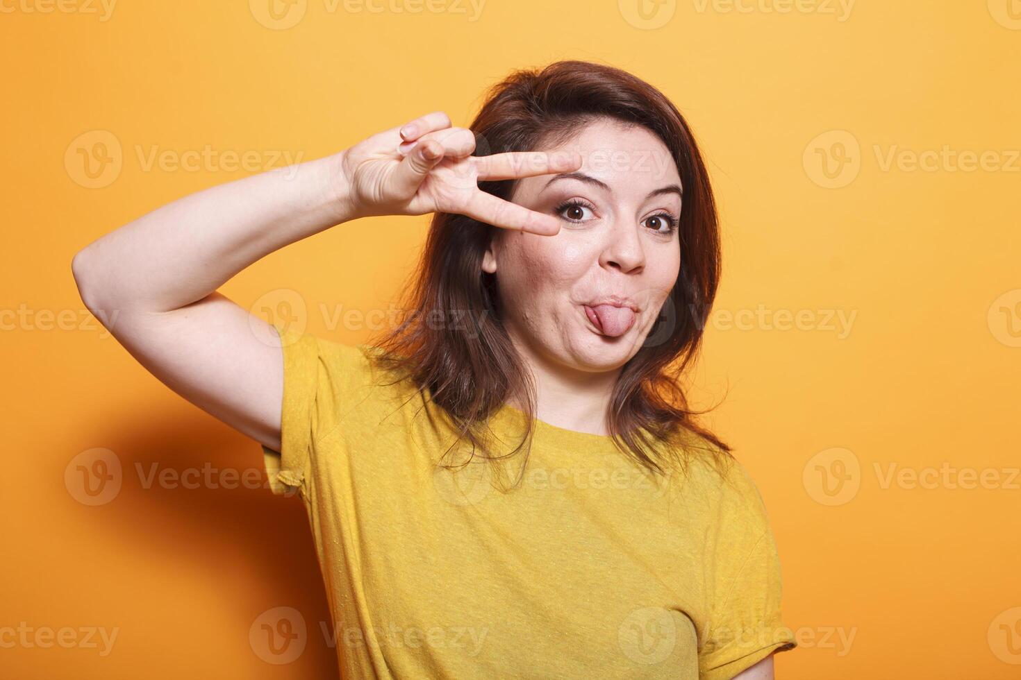 Portrait of caucasian female individual doing peace sign and having her tongue out, showing victory gesture with fingers. Advertising peaceful success symbol and fooling around on studio camera. photo