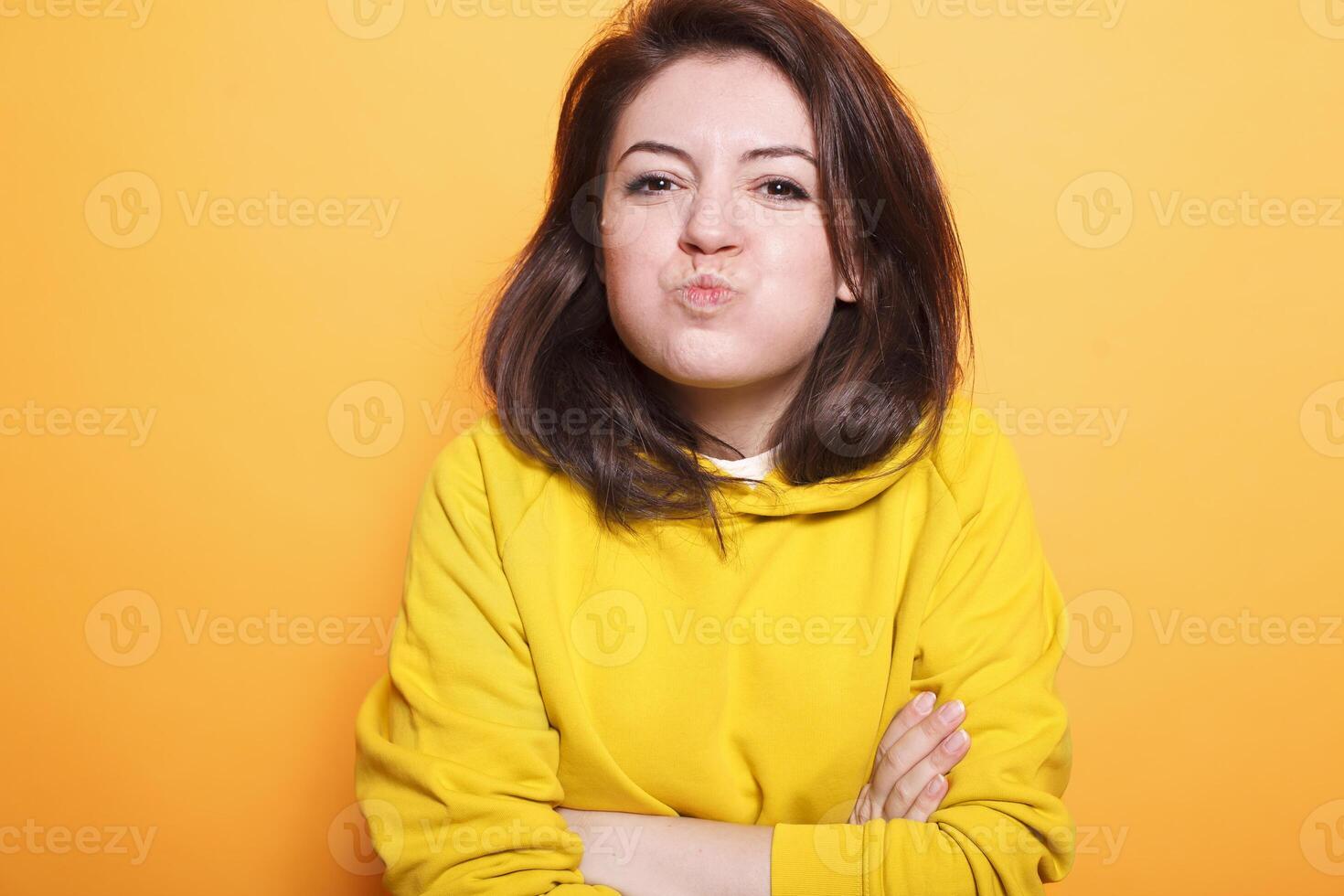 Confident beauty model with brunette hair makes a kissy face on camera while posing carefree in the studio. Portrait of young female adult with arms crossed making a goofy face. photo