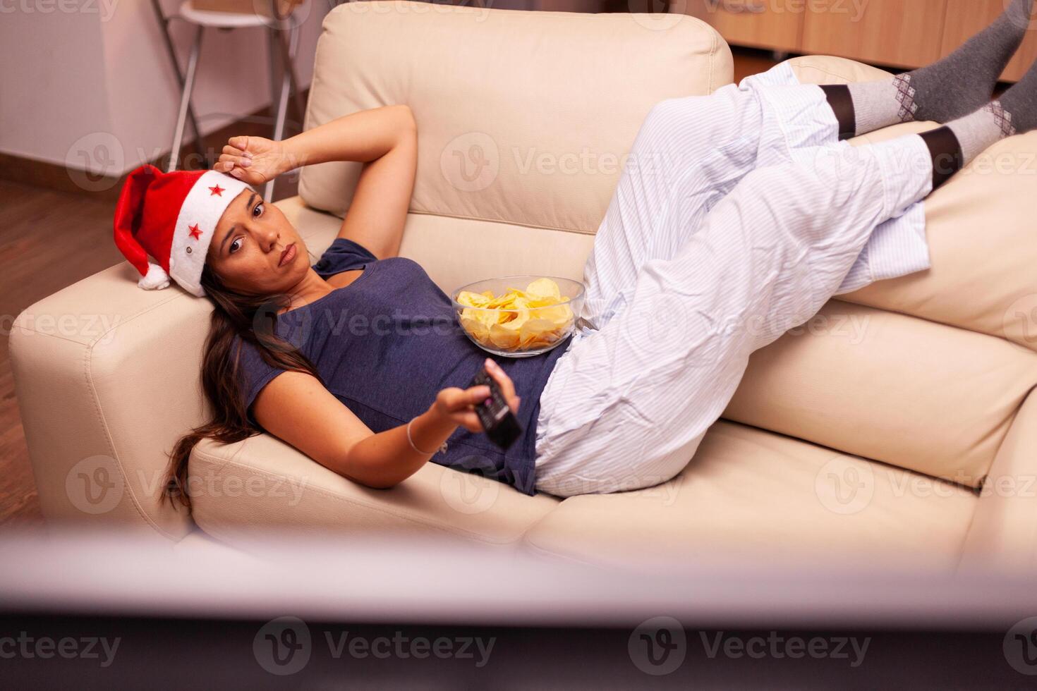 Relaxed woman resting on couch watching christmas movie series on television celebrating christmastime in x-mas decorated kitchen. Girl wearing santa hat enjoying winter season at home photo