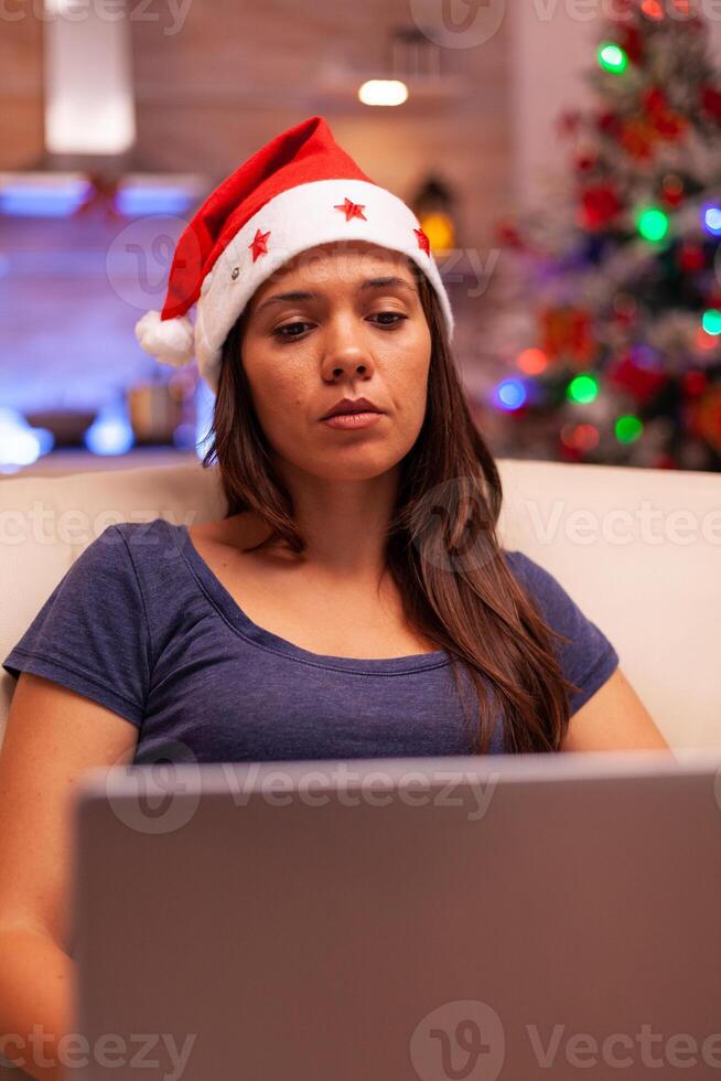 Girl looking at laptop screen reading business email during christmas holiday sitting comfortable on sofa in xmas decorated kitchen. Woman adult browsing on social media. Winter season photo