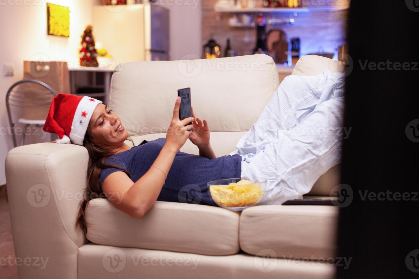 Woman lying on sofa browsing on social media using smartphone during christmastime in xmas decorated kitchen. Girl enjoying winter holiday celebrating christmas season at home photo