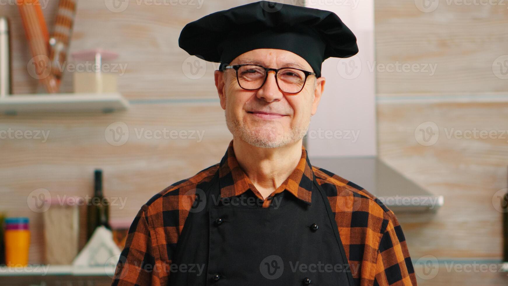 Senior baker smiling looking at camera in kitchen with food ingredients on table for baking. Retired elderly chef with bonete ready to cook homemade bread, cakes and pasta with wheat flour and eggs photo