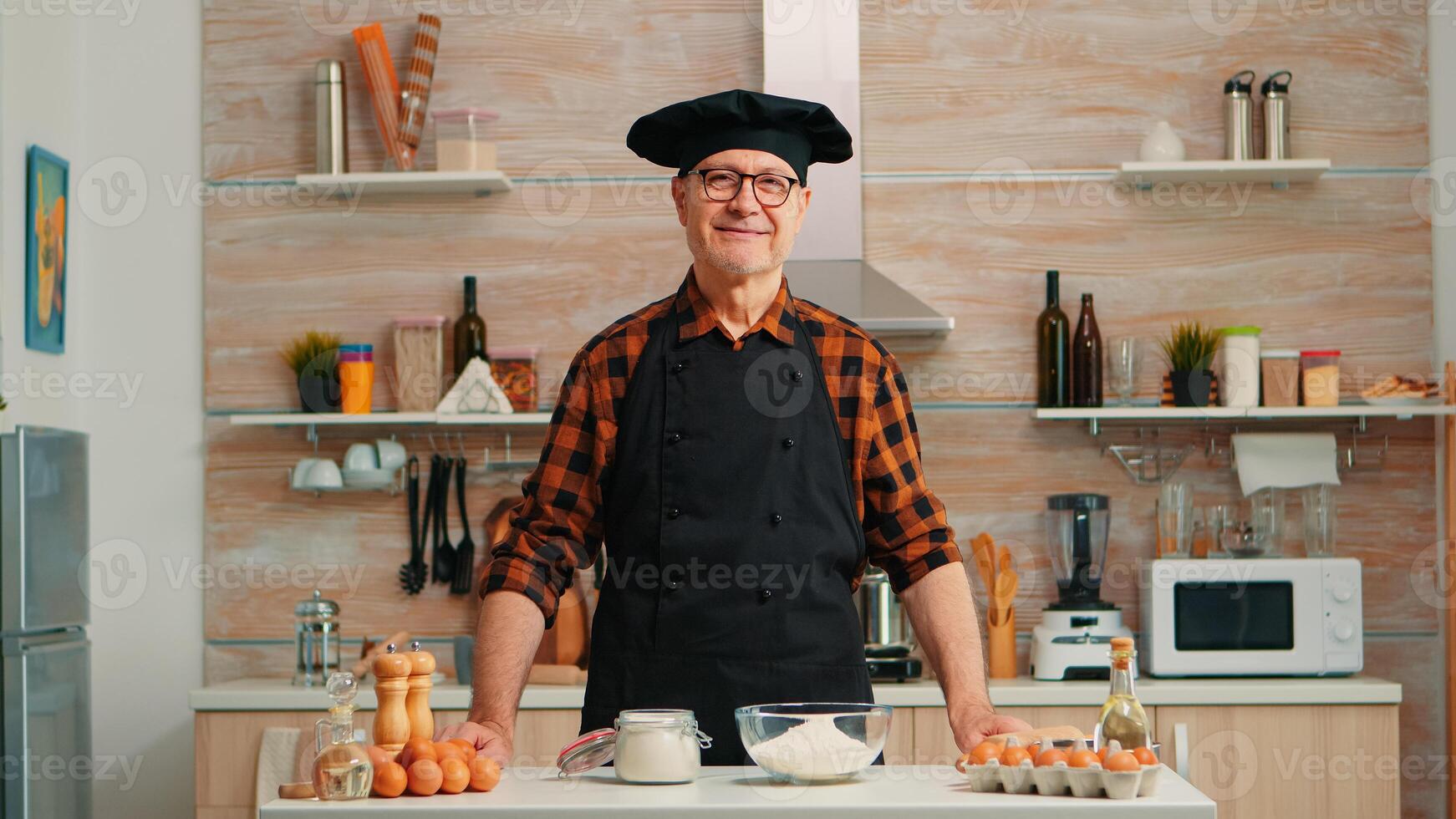 Portrait of chef wearing bonete looking at camera and smiling. Retired elderly baker in kitchen uniform preparing pastry ingredients on wooden table ready to cook homemade tasty bread, cakes and pasta photo