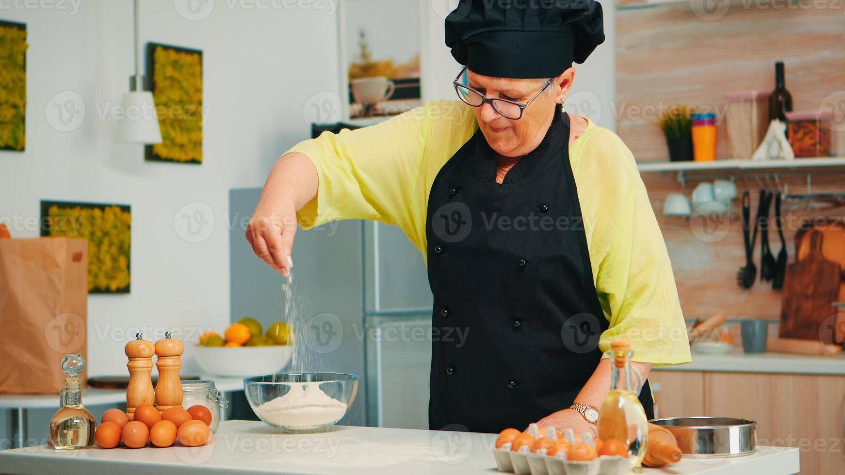 Baker spreading flour on wooden table at home in modern kitchen wearing apron and bonete. Happy elderly chef with uniform sprinkling, sieving sifting raw ingredients by hand baking homemade pizza photo