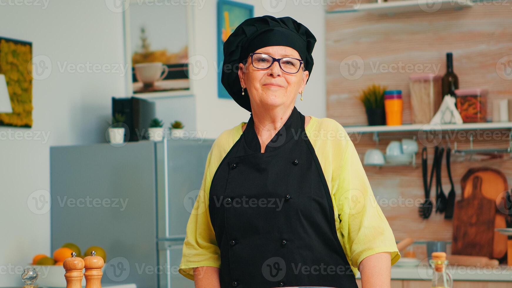 Old lady wearing chef apron and bonete in home kitchen with flour on table for baking looking at camera and smile. Retired elderly baker preparing bakery ingredients to cook homemade bread and cakes photo