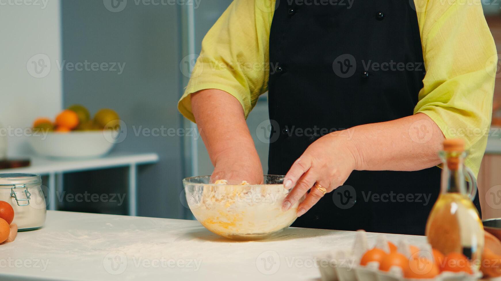 Chef senior woman mixing by hand bread ingredients in home kitchen in glass bowl. Retired elderly baker with bonete kneading cracked fresh eggs with wheat flour baking homemade cake and bread photo