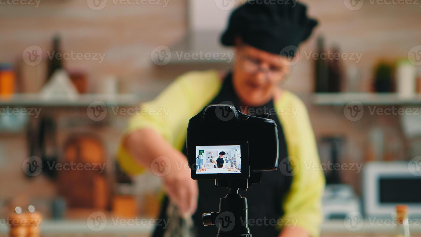 Old lady baker presenting food recipe on cooking podcast from kitchen. Retired blogger chef influencer using internet technology communicating, shooting blogging on social media with digital equipment photo