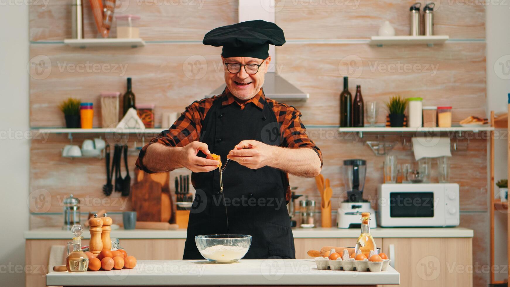 Experienced baker man craking eggs for baking wearing apron enjoying hobby. Retired elderly chef with bonete mixing by hand, kneading in glass bowl pastry ingredients preparing homemade cake photo